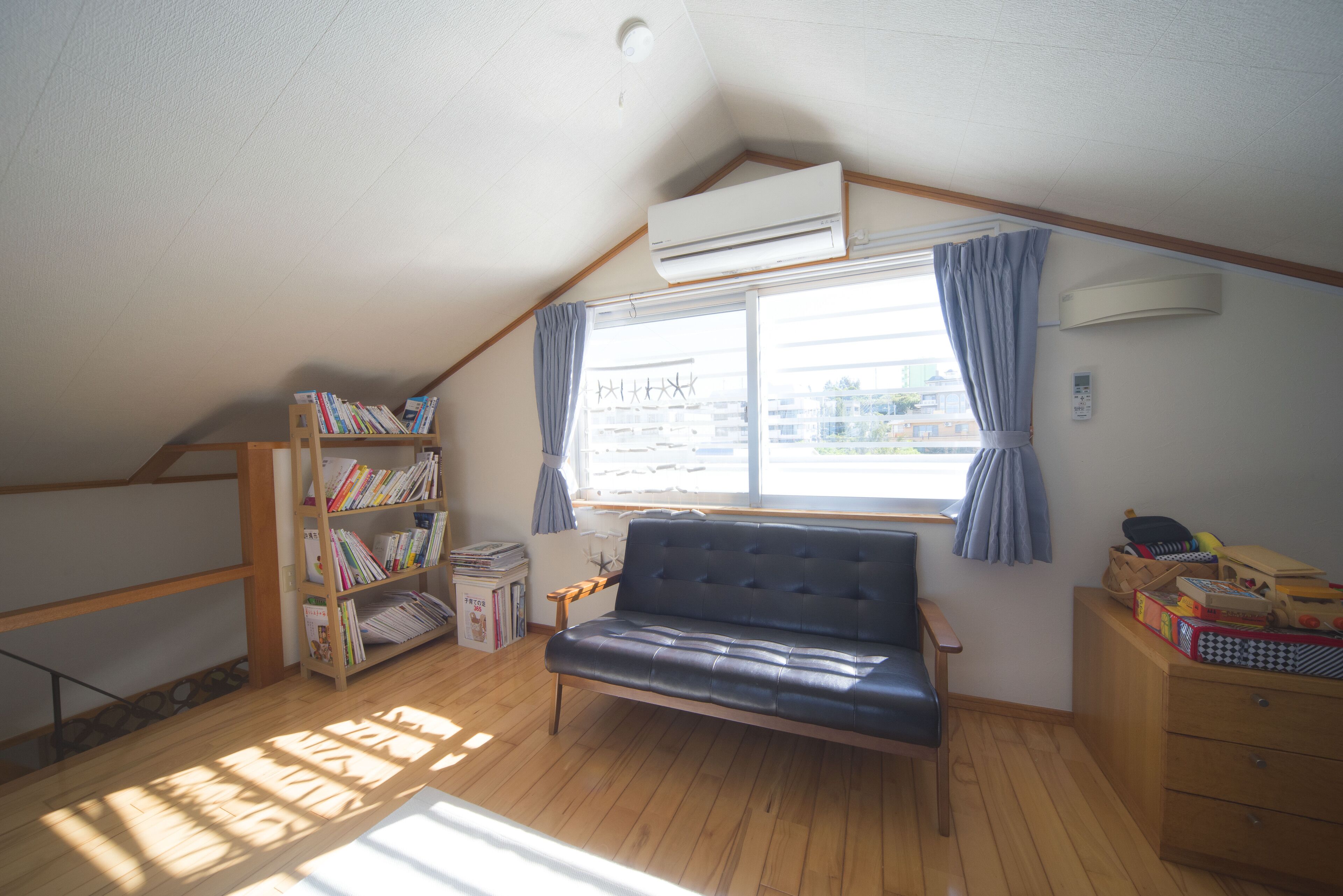 A room with a sloped ceiling, a large window with curtains, a sofa, a bookshelf, and a wooden dresser.