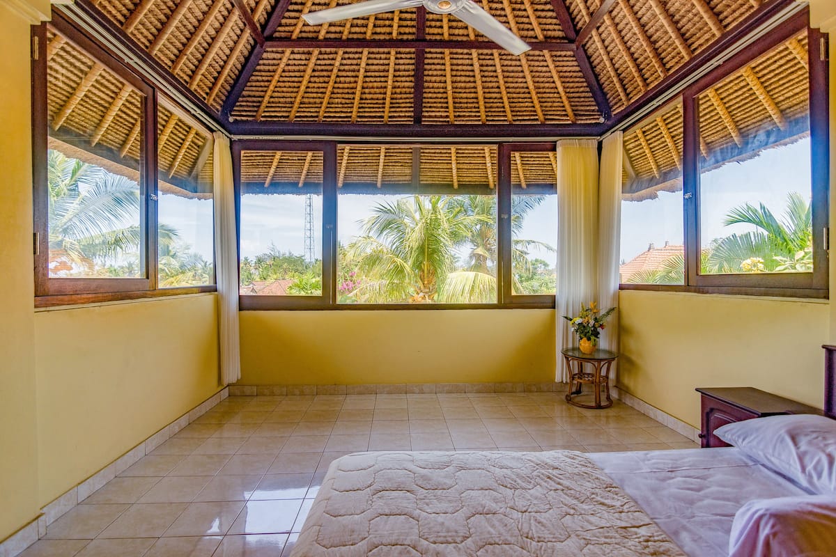 A room with a bed, a ceiling fan, large windows, and a view of palm trees.
