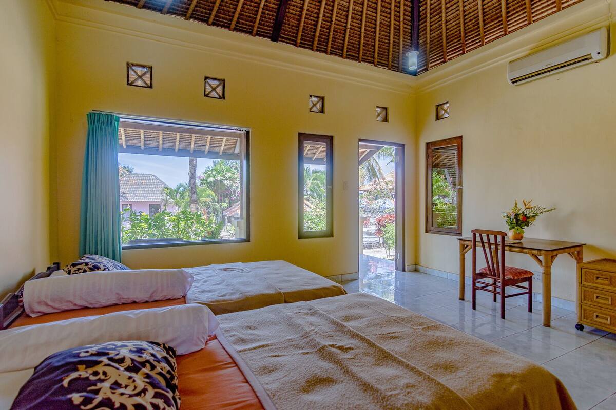 A hotel room with two beds, a desk, and a chair. There are windows with views of greenery and a building outside.
