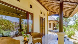 A covered outdoor area with wicker furniture, a potted plant, and a view of a building with a thatched roof. - Villa Sari Cucukan (Gianyar)