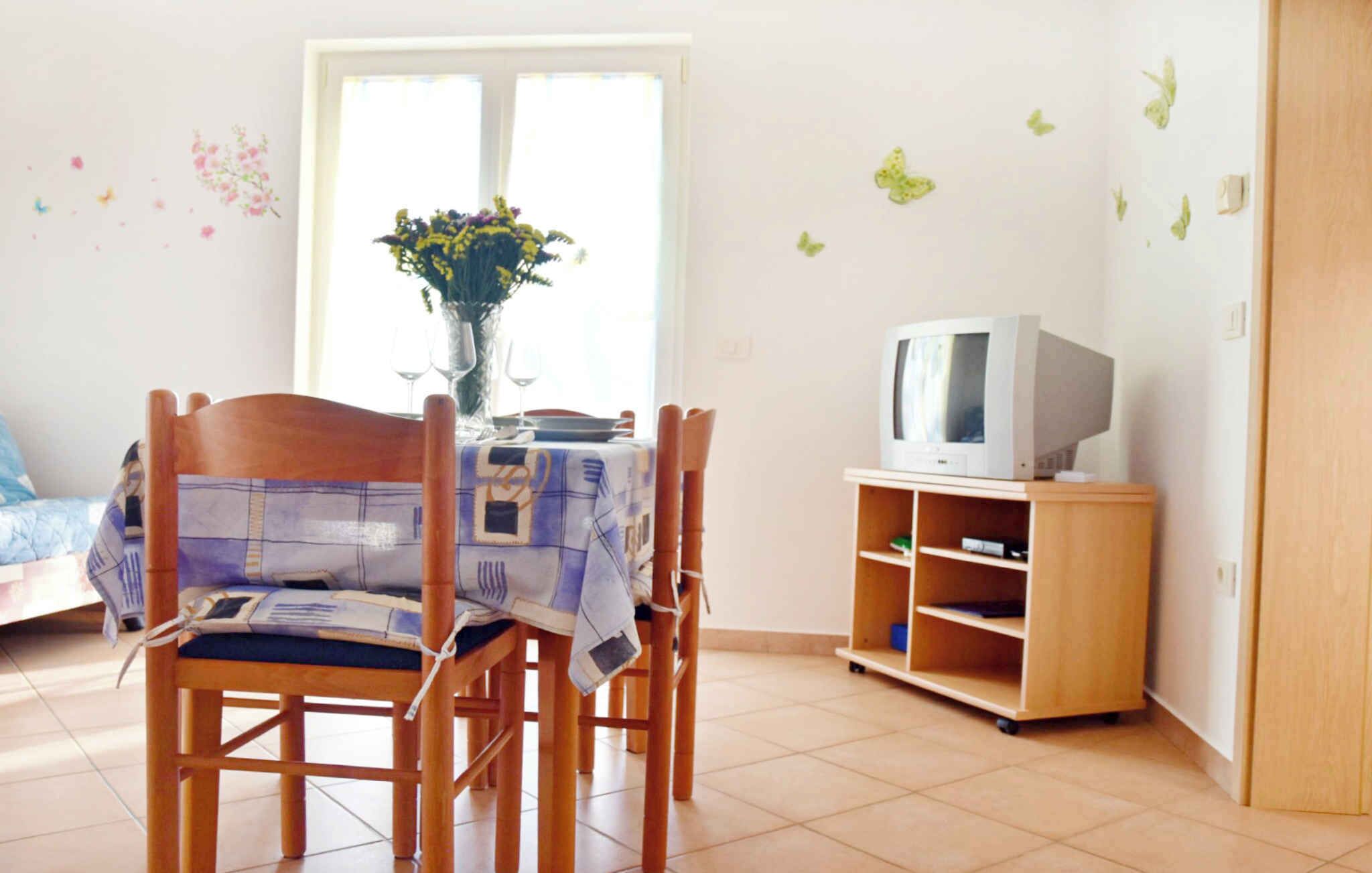 A room with a dining table set for four, a television on a wooden stand, and a window with a view of the outside.