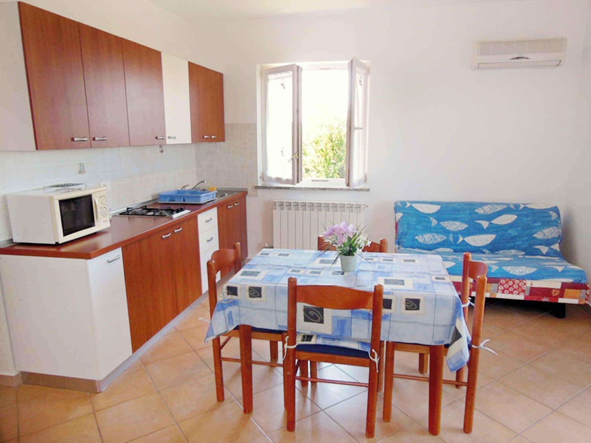 A compact kitchen with wooden cabinets, a microwave, and a dining table with chairs. There is a window with an open shutter and a radiator below.
