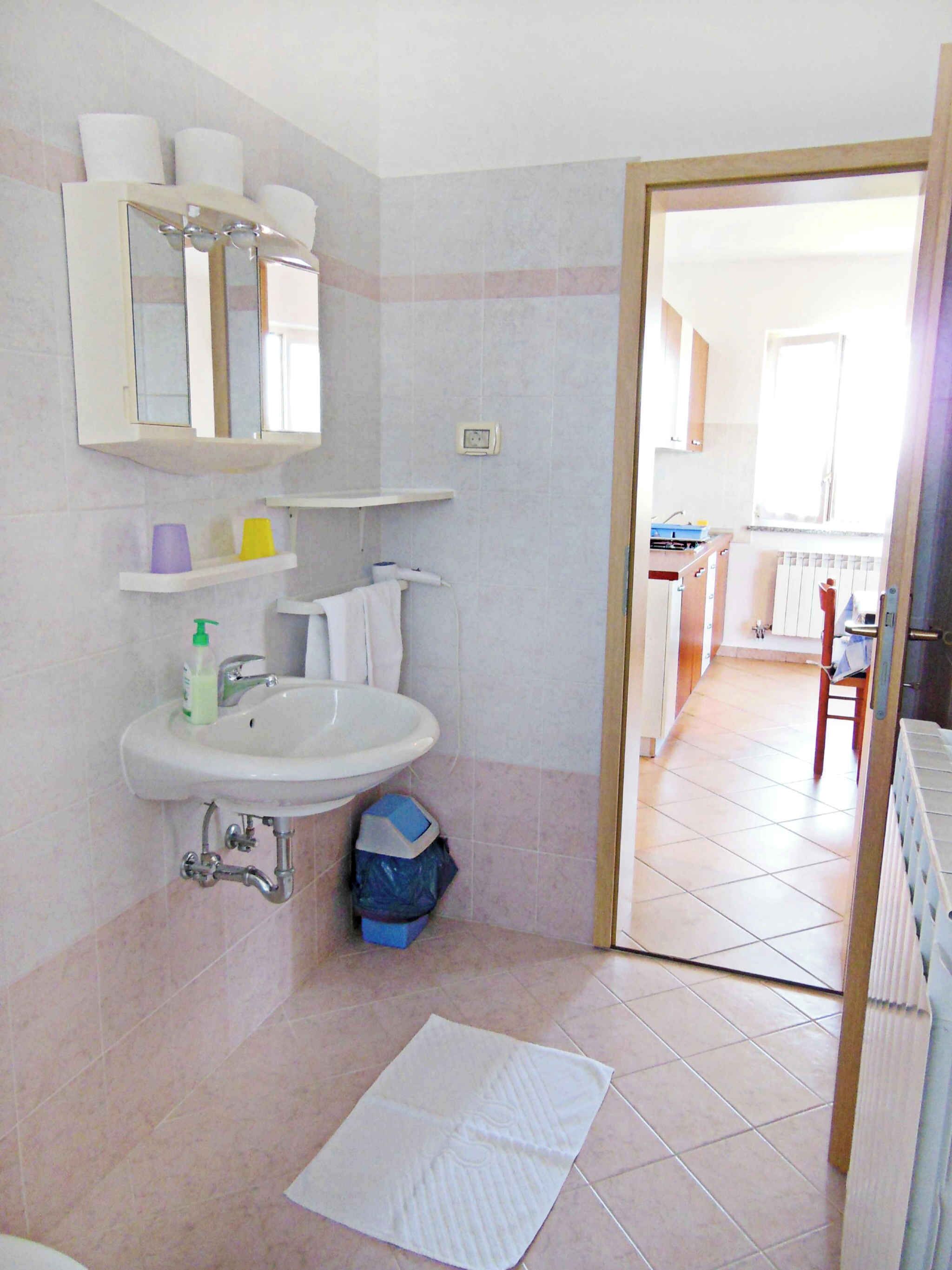 A bathroom with a sink, mirror, and shelves. There is a door leading to a kitchen area.