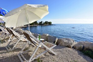 A beachside setting with wooden lounge chairs and a white umbrella, a bottle of beer, and a rocky shoreline with calm water. - Holiday apartment with balcony, air conditioning and wifi (Zambratija)