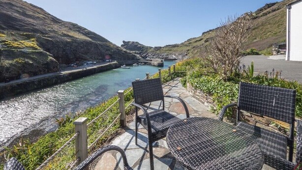 A riverside patio with wicker furniture overlooking a river, surrounded by greenery and hills. - A beautiful harbourside four-bedroom cottage with harbour views (Boscastle)