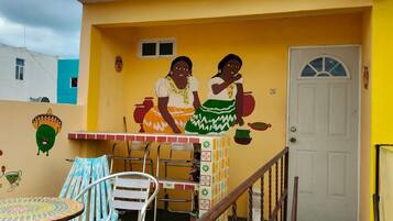 A building with a yellow exterior, a white door, and a mural of two women. There is a table with chairs and a small table with a patterned tablecloth.