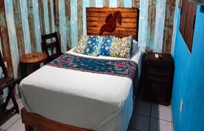 A bed with a floral bedspread, a wooden headboard with a horse carving, a small round table, and a nightstand with a lamp. - Cancun Guest House 5 (Cancún)