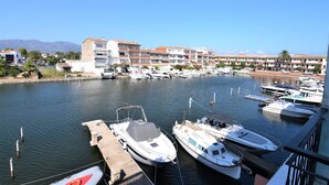 Un port de plaisance où sont amarrés plusieurs bateaux, un immeuble doté de nombreux balcons et une vue dégagée sur l’eau et les montagnes lointaines.