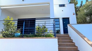 Uma casa moderna de dois andares com telhado plano, porta azul e escadaria que leva à entrada.