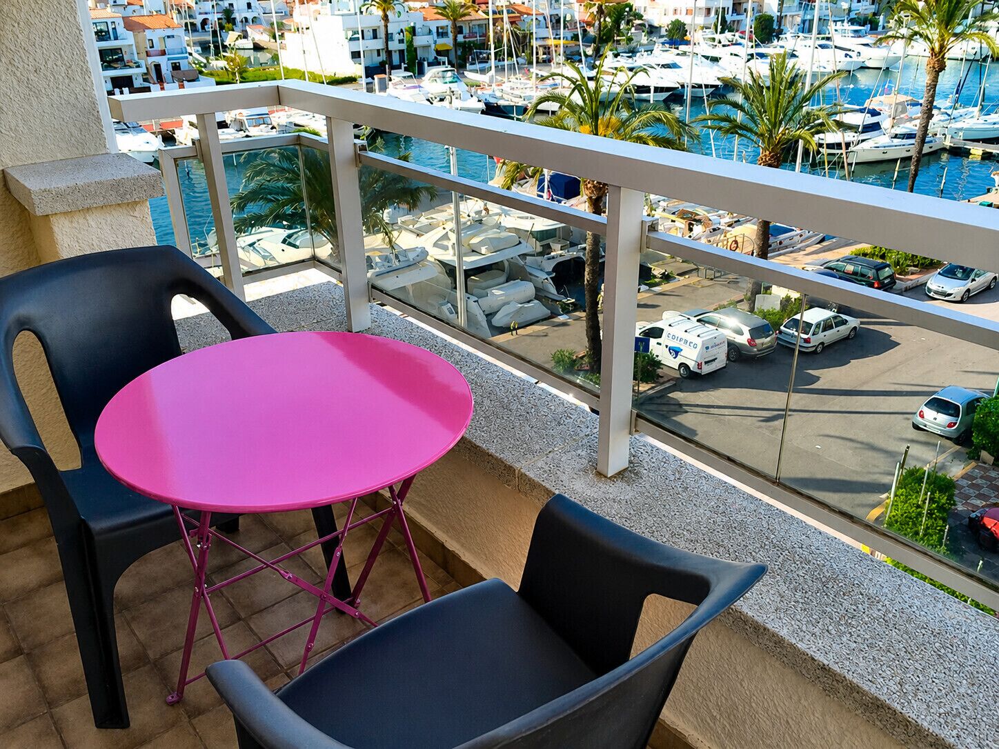 Un balcón con una mesa rosa y dos sillas, con vista a un puerto deportivo con barcos y un estacionamiento.