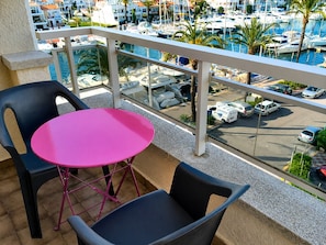 Un balcón con una mesa rosa y dos sillas, con vista a un puerto deportivo con barcos y un estacionamiento.