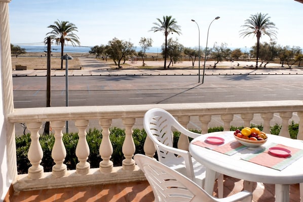 A balcony with a table and chairs, a bowl of fruit, and a view of a road with palm trees and a distant body of water.