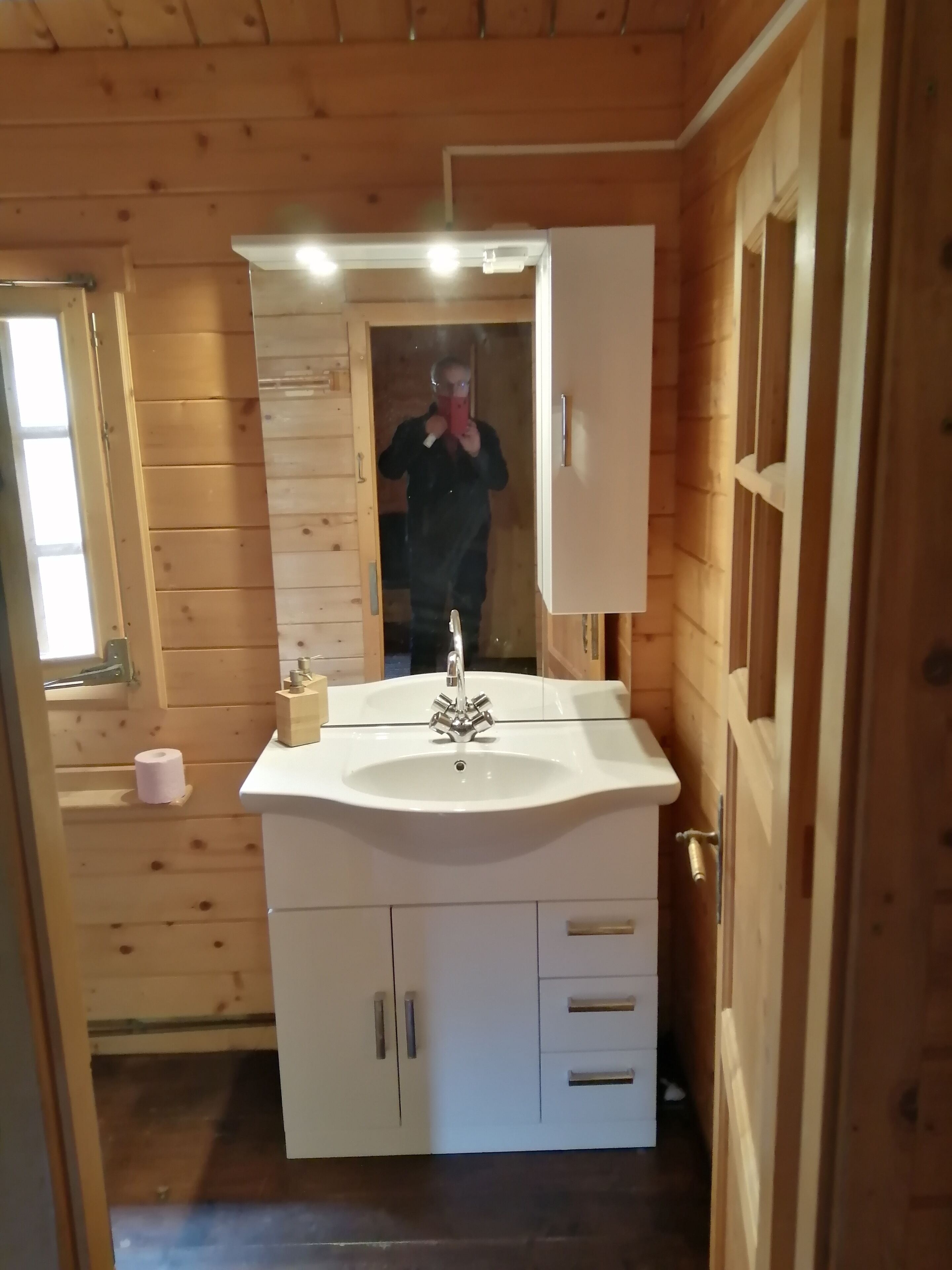 Ett badrum i en stuga med träpanel, ett vitt handfat, en spegel och en trädörr.