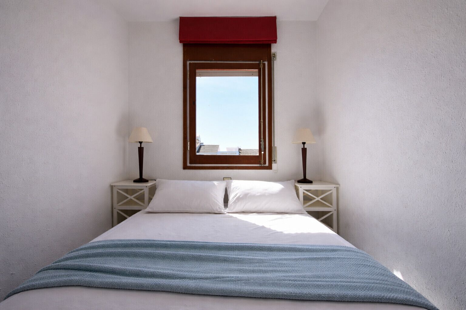 Une petite pièce comprenant un lit, deux tables de chevet avec des lampes, une fenêtre avec un store rouge et une vue sur les bâtiments environnants.