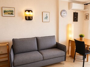 A modern living room with a grey sofa, a wooden dining table with chairs, a wall-mounted clock, and framed artwork.