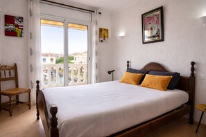 A bedroom with a bed, a chair, a window with a view, and a painting on the wall.