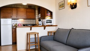 Eine kompakte Küche mit Holzschränken, einem weißen Kühlschrank, einer Mikrowelle und einer kleinen Arbeitsfläche. Im Wohnbereich befinden sich zwei Hocker und ein graues Sofa.