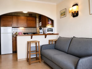 Une cuisine compacte avec des meubles en bois, un réfrigérateur blanc, un four à micro-ondes et un petit plan de travail. On y trouve deux tabourets et un canapé gris dans l’espace salon.