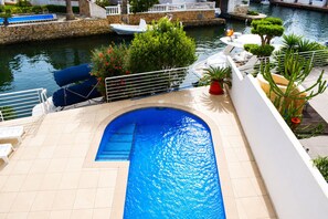 A swimming pool with a stone border, a boat docked nearby, and a view of a canal with boats. - 306-Magnificent modern house with pool private and mooring (Empuriabrava)