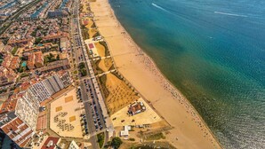 Vue aérienne d’une ville côtière avec une vaste plage, un port de plaisance et une zone urbaine dense, avec des immeubles et des routes.