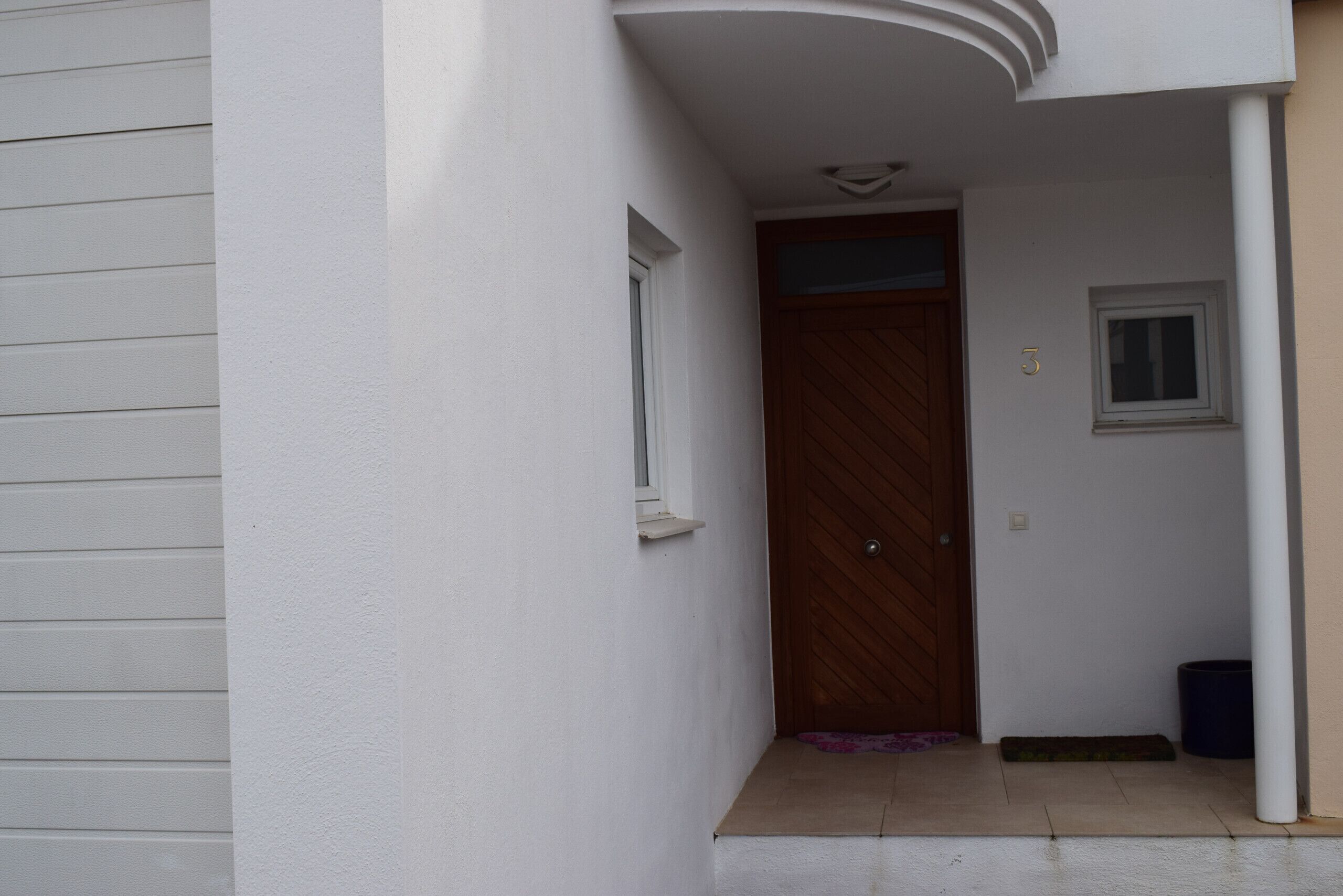 A white exterior wall with a wooden door, a small window, and a trash bin.