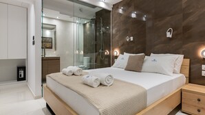 A modern hotel room with a large bed, a glass shower enclosure, a wooden nightstand, and a built-in wardrobe. - Rhodes Skyline Suite - 2 Bedroom with OutDoor  jacuzzi and Sea view (Rhodes)