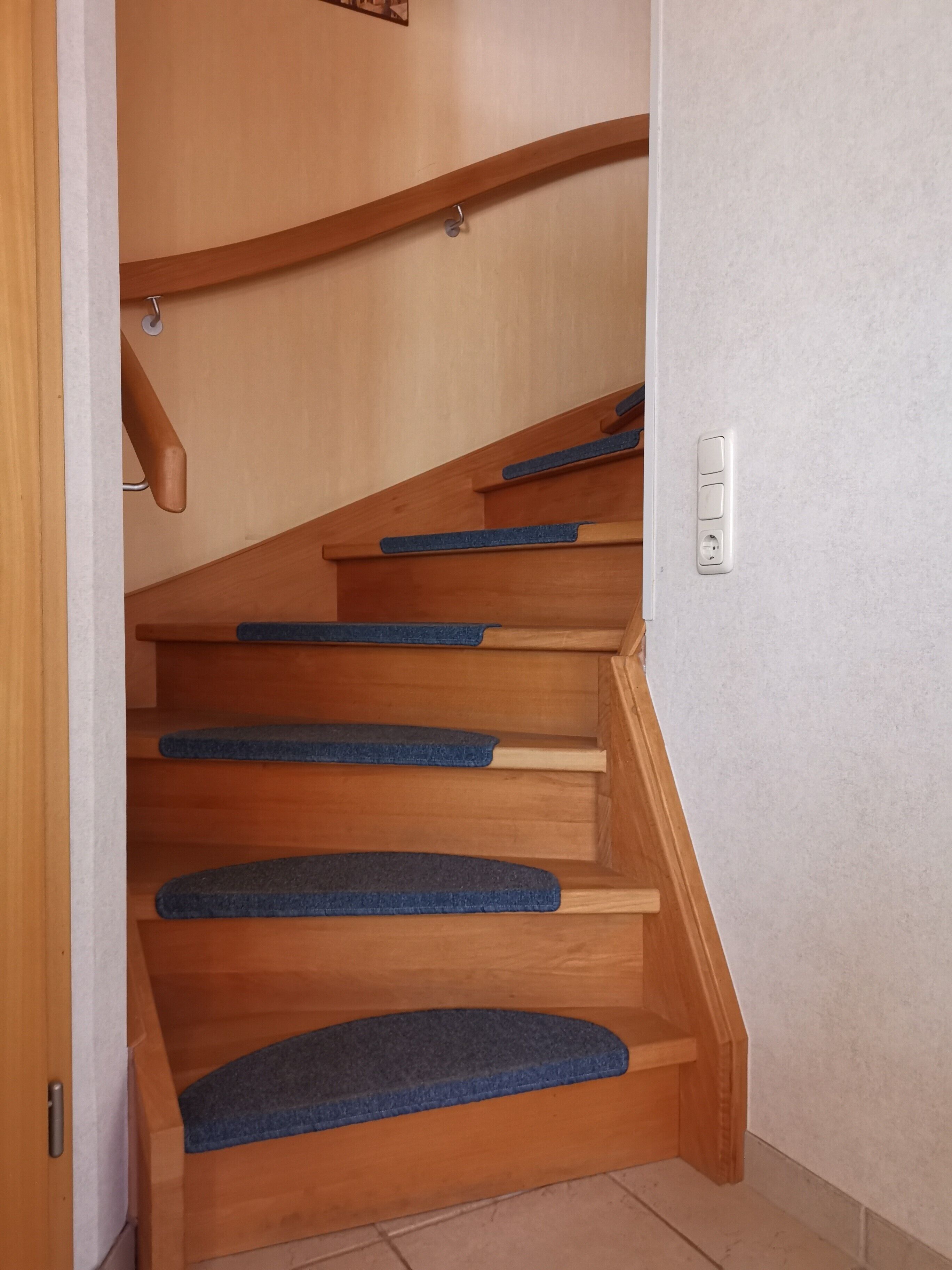 Een houten trap met blauw beklede treden en een witte muur met een lichtschakelaar.