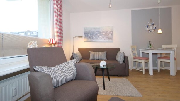 Ein Wohnzimmer mit einer Couch, einem Sessel und einem kleinen Tisch mit Lampe. Ein Fenster mit Vorhängen, ein Bild an der Wand und ein Tisch mit Stühlen sind ebenfalls zu sehen.