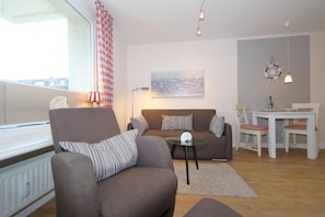 Un salón con sofá, sillón y una mesita con lámpara. Hay una ventana con cortinas, un cuadro en la pared y una mesa con sillas.