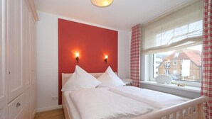 Ein Schlafzimmer mit Bett, weißen Wänden, einer roten Akzentwand, zwei Wandleuchten, einem Fenster mit Vorhängen und Blick auf Gebäude.