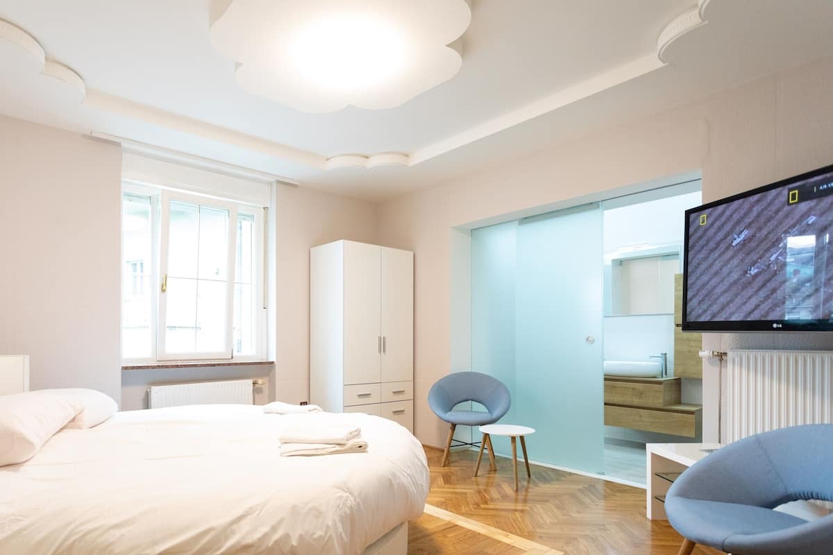 Ein modernes Hotelzimmer mit einem großen Bett, einem Schrank, einer Sitzecke mit Stuhl und Tisch sowie einem an der Wand befestigten Fernseher.