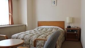 Een hotelkamer met een bed, een stoel, een klein tafeltje, een nachtkastje met een lamp, en een lijstje aan de muur.