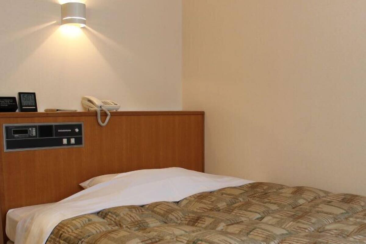 Hotelkamer met een houten hoofdeinde, nachtkastje met telefoon en wekker, en bed met een geruit plaiddeken.