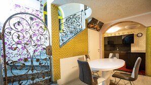 Un coin repas avec une table ronde et deux chaises, un escalier agrémenté de rampes en métal décoratives, et un espace cuisine avec une horloge au mur.