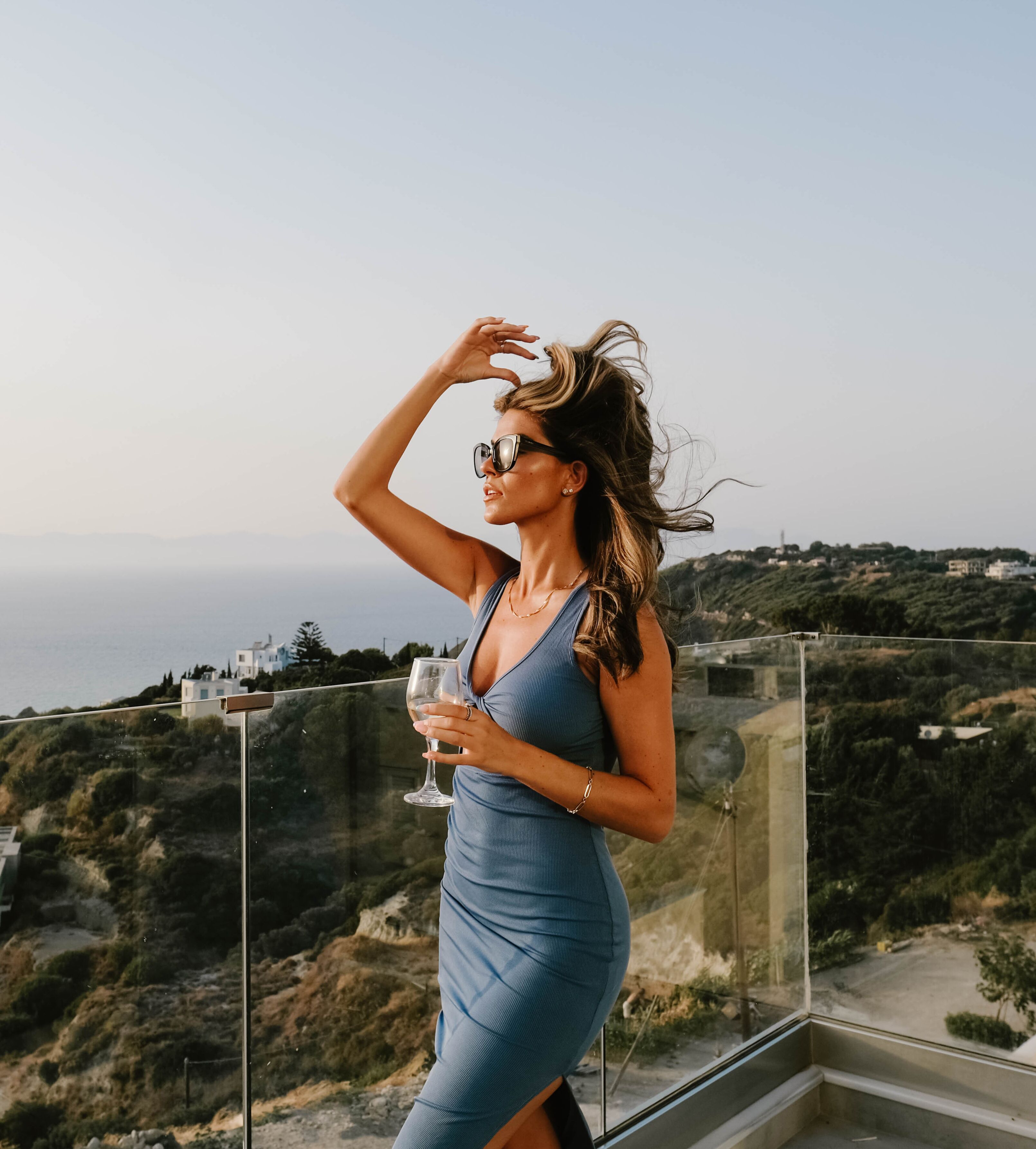 A woman in a blue dress standing by a glass railing overlooking a coastal landscape.