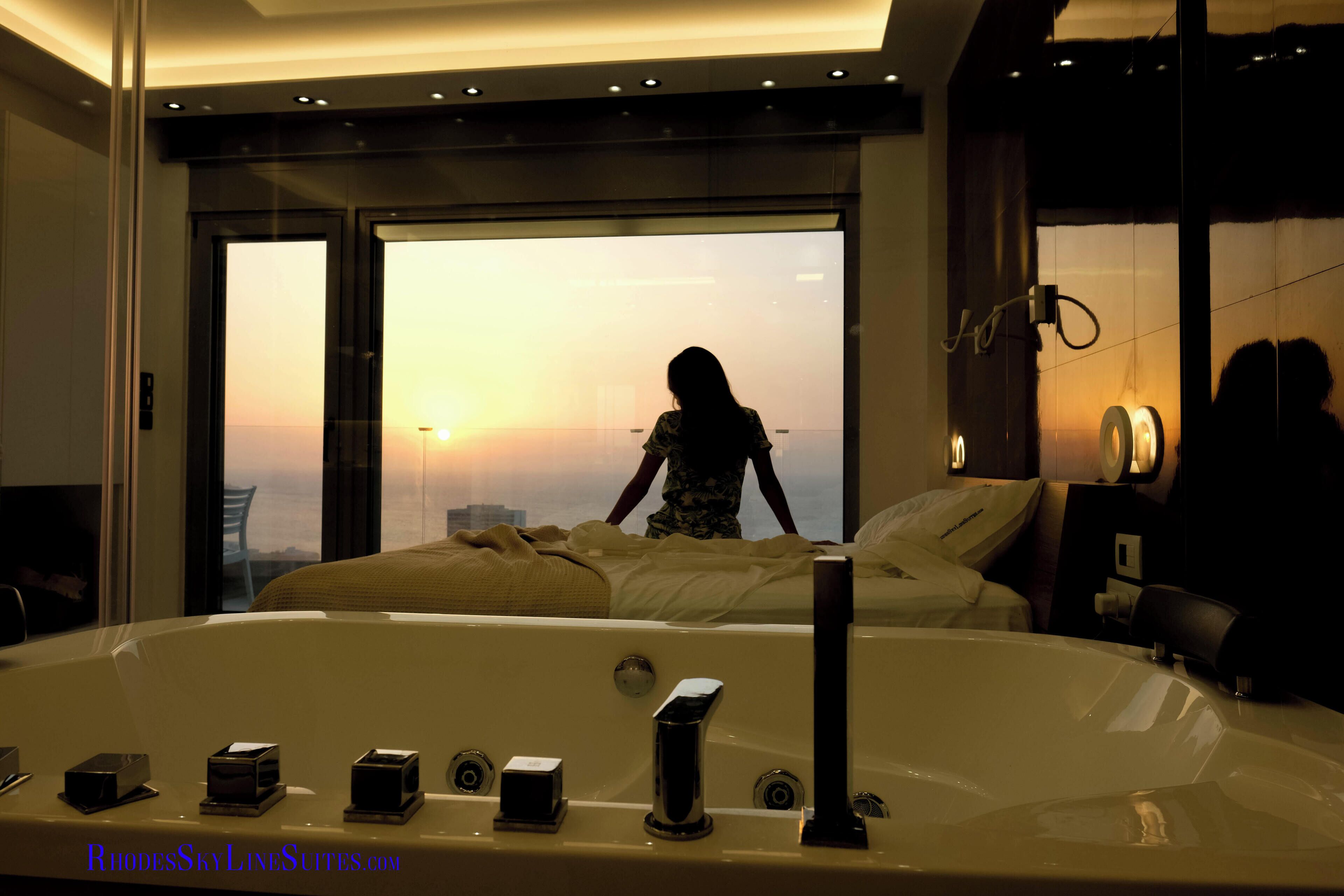 Une chambre spacieuse avec un grand lit, une salle de bain séparée avec une baignoire, et une vue sur le paysage urbain au coucher du soleil.