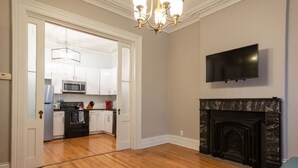 A living room with a fireplace, a television mounted on the wall, and a kitchen visible through an open door.
