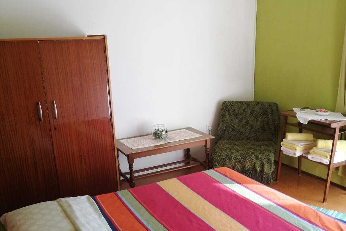 A bedroom with a bed, a wooden wardrobe, a small table with a glass container, a chair, and a shelf with towels.