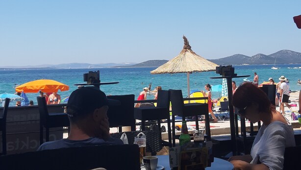 En restaurang vid stranden med uteservering, parasoller och utsikt över havet och bergen.