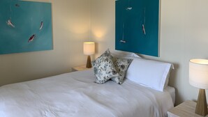 Un lit bien fait, avec du linge de lit blanc, un coussin à motifs floraux et deux tableaux représentant des voiliers, accrochés au mur.