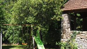 Un terrain de jeux avec des balançoires et un toboggan, entouré d’arbres et d’un mur de pierre.