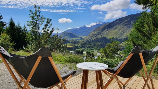 Two black folding chairs with a small round table between them, set on a wooden deck overlooking a mountainous landscape with a clear sky. - Entire 2 bedroom detached chalet, in private woodland with valley views. (Arthurs Point)
