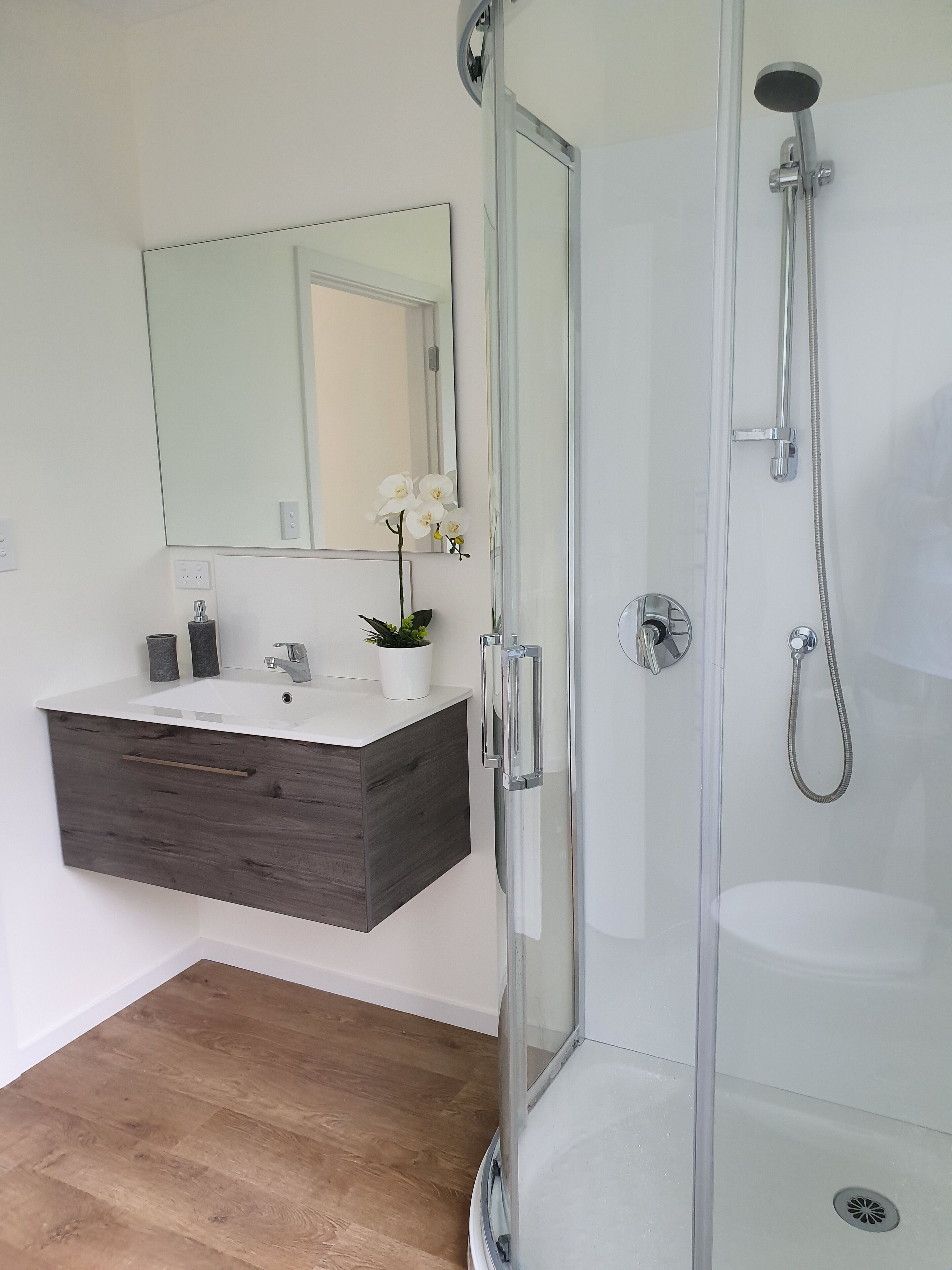 A modern bathroom with a glass-enclosed shower, a wooden vanity with a mirror, and a potted orchid.