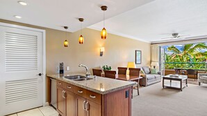 Une cuisine moderne dotée d’un îlot central, d’un coin repas avec un canapé et des fauteuils, et offrant une vue sur des plantes tropicales grâce à une porte-fenêtre coulissante.