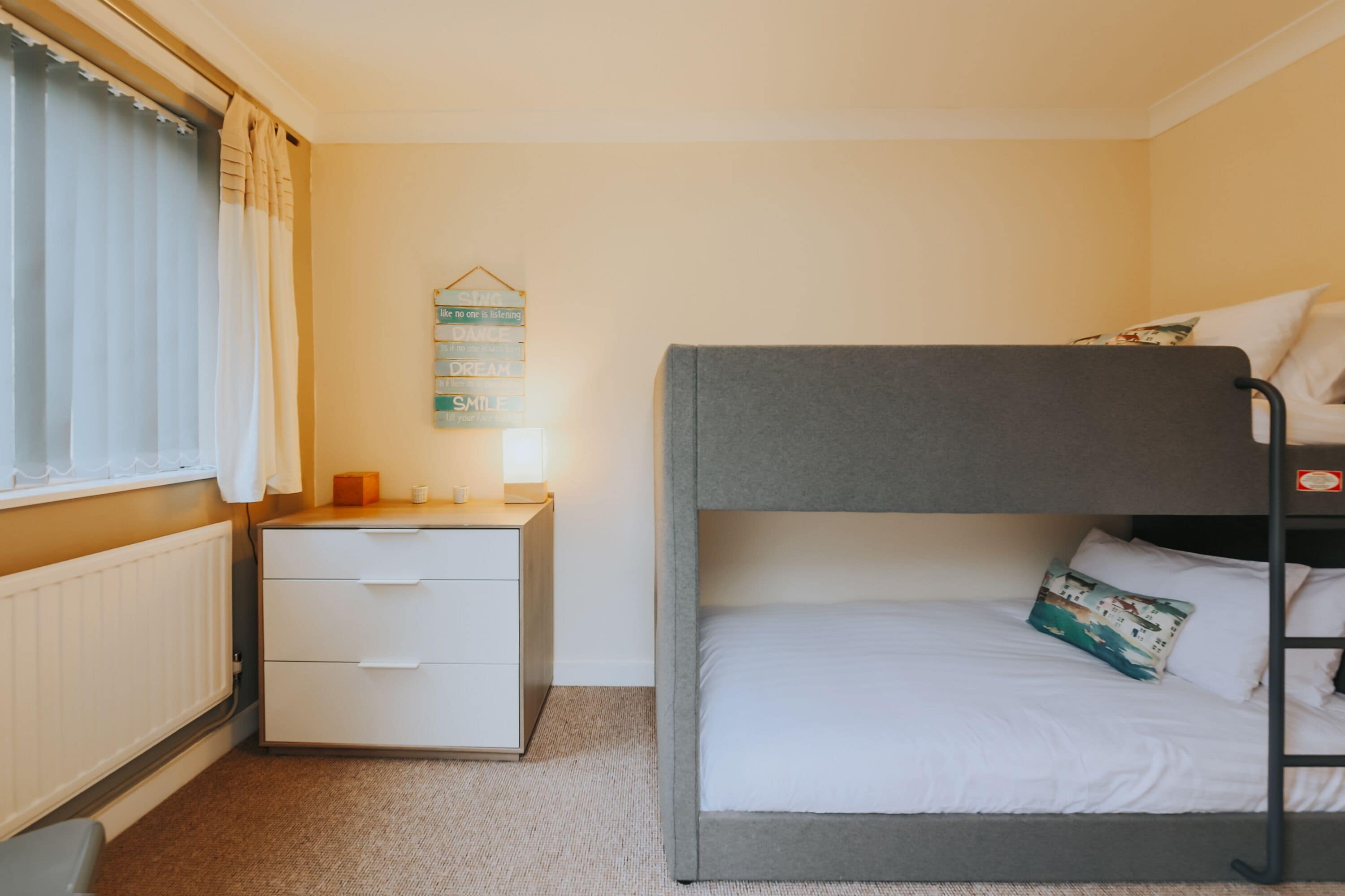 Uma cama beliche com colchão e almofadas brancos, uma mesinha de cabeceira com abajur, um penteou branco e uma janela com cortinas.