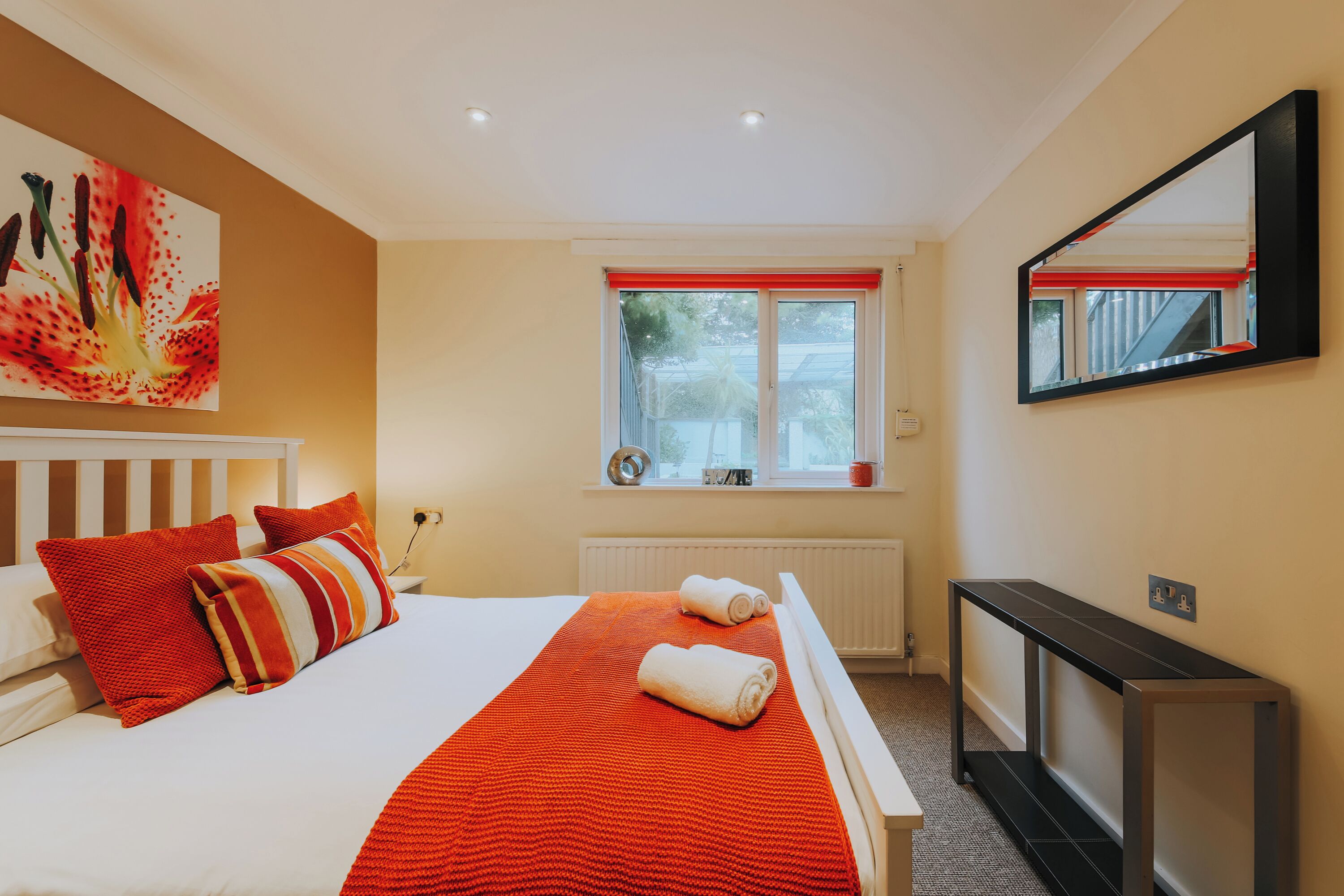 A hotel room with a bed, a large window with red blinds, a bedside table, and a mirror on the wall.