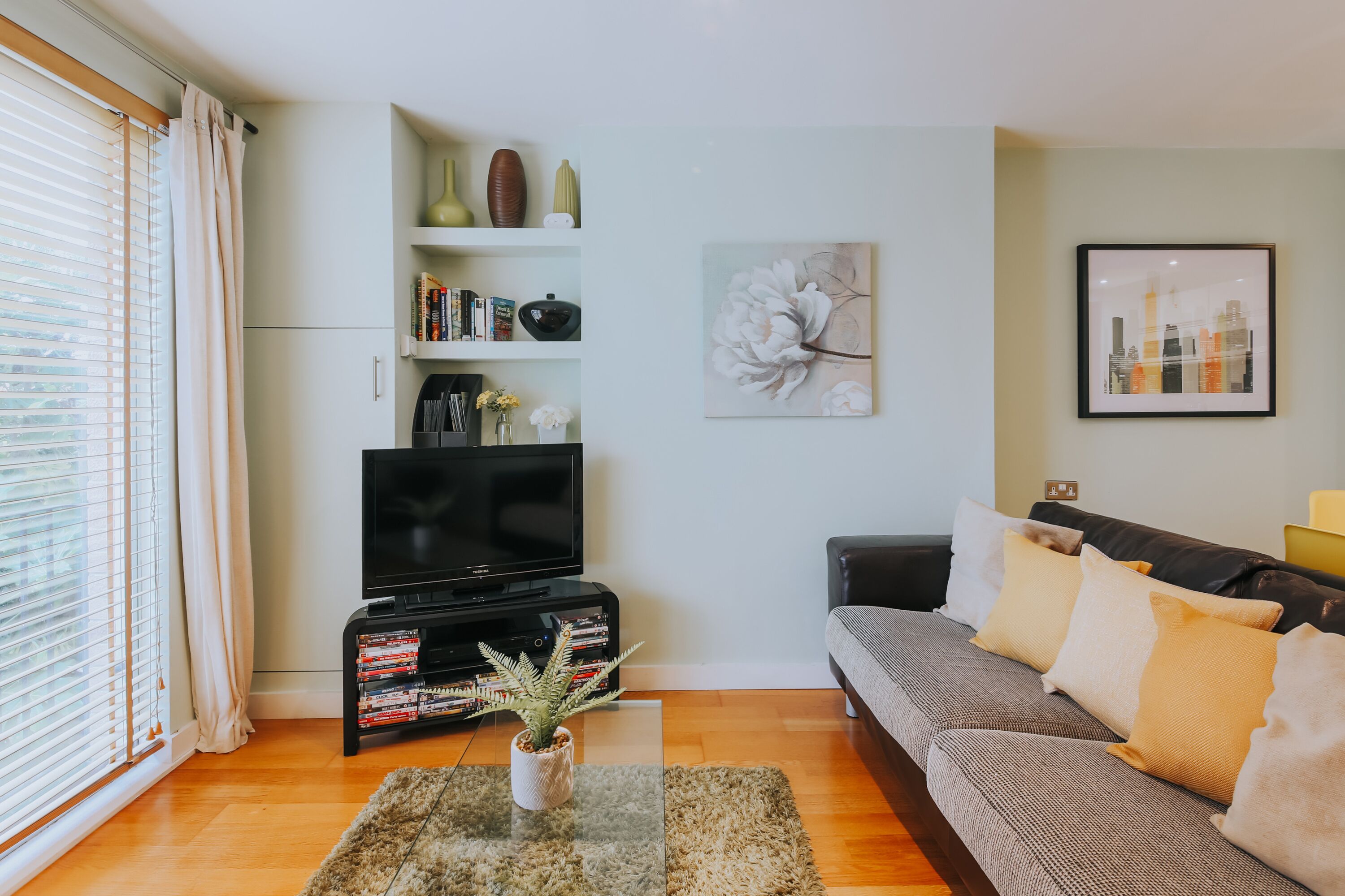 A living room with a flat-screen TV, a bookshelf, a sofa, and a window with blinds.