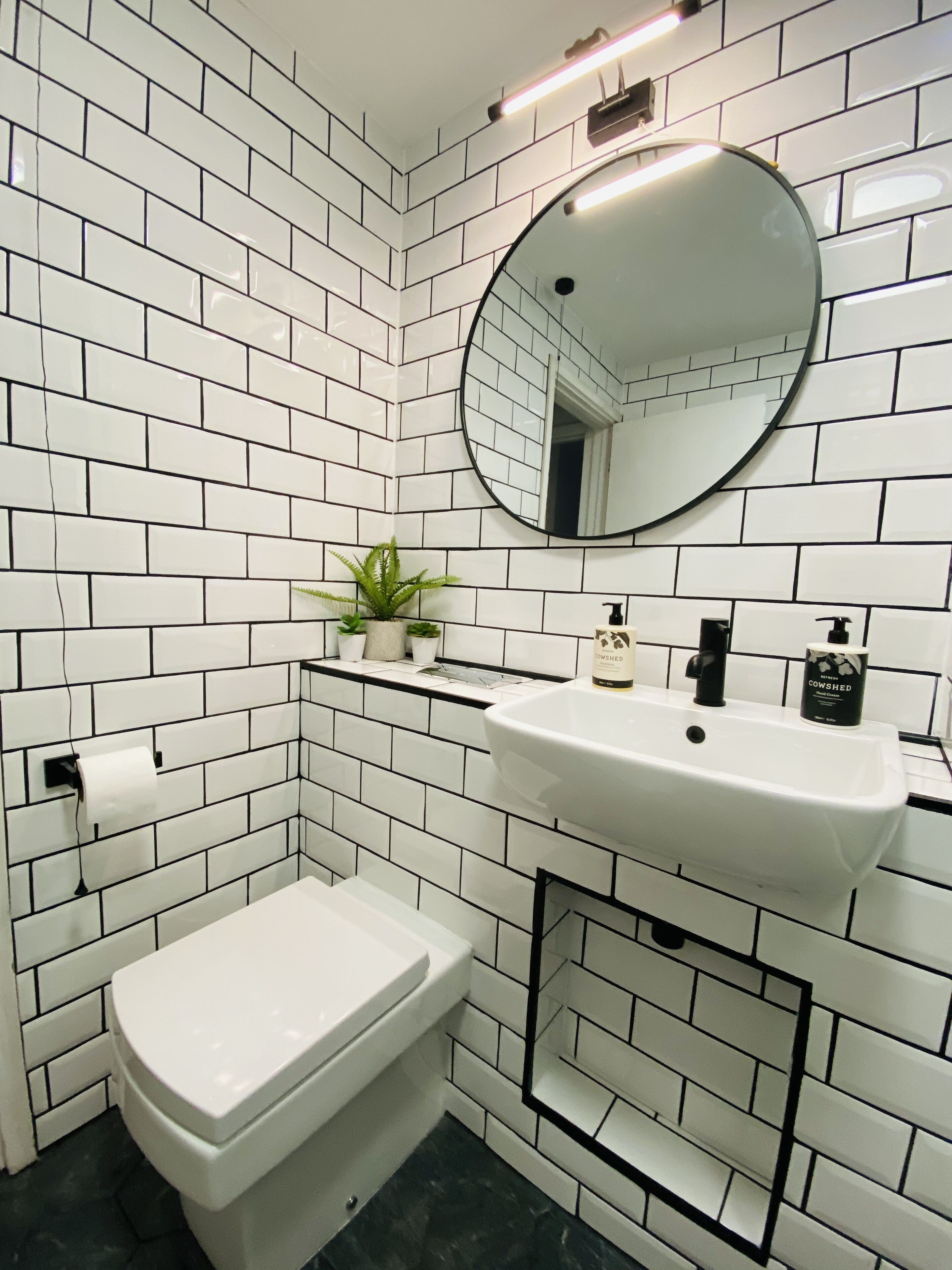 Un baño con azulejos blancos tipo metro, un lavamanos blanco, un inodoro, un espejo y una pequeña planta en maceta.