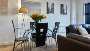 A modern living room with a glass dining table, black chairs, a brown leather sofa, and a vase of flowers. There are two framed cityscape paintings on the wall. - Clarence Court (Newcastle-upon-Tyne)