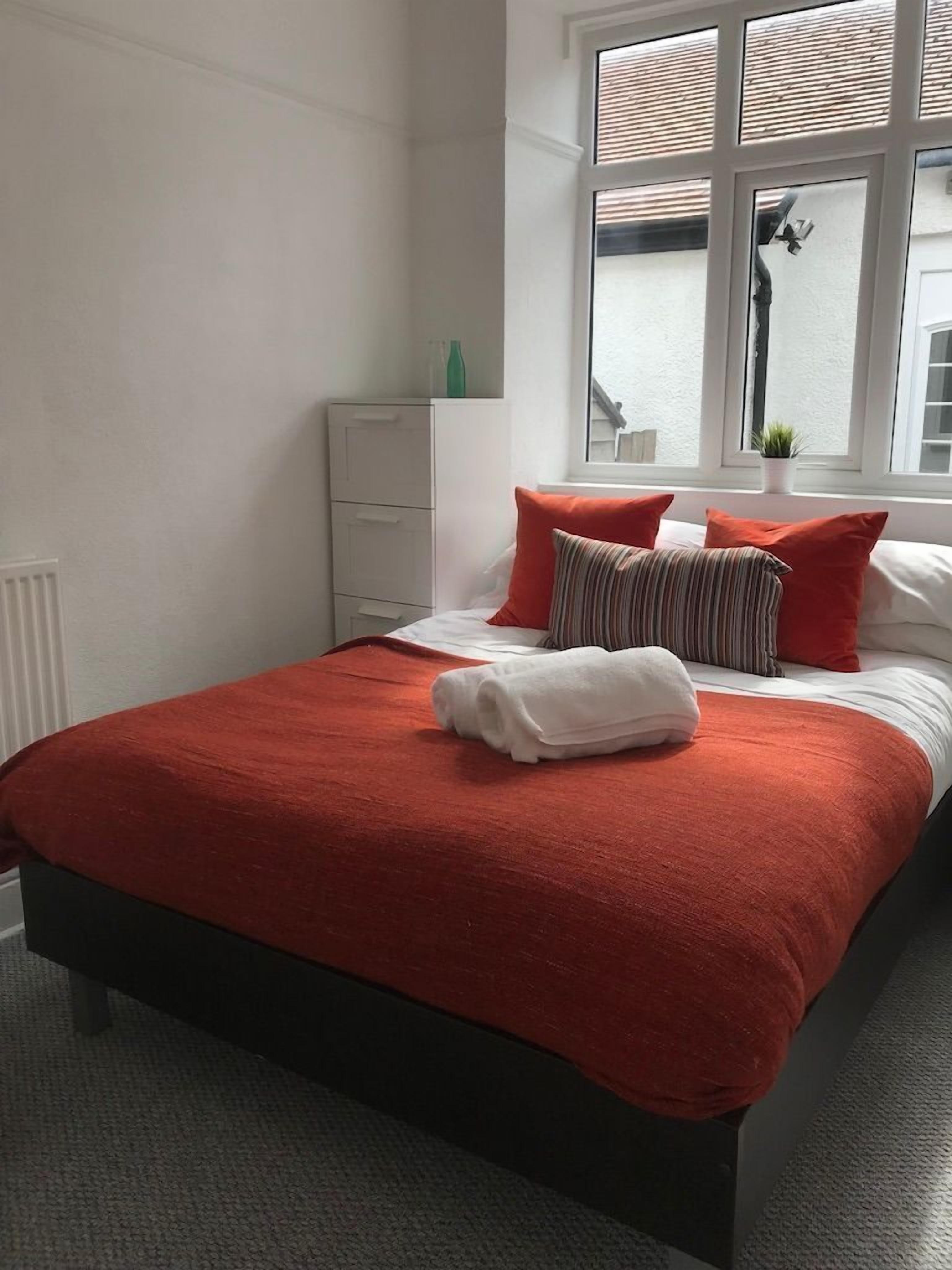 Een net opgemaakt bed met een rode deken en kussens, een witte nachtkastje en een raam met jaloezieën.